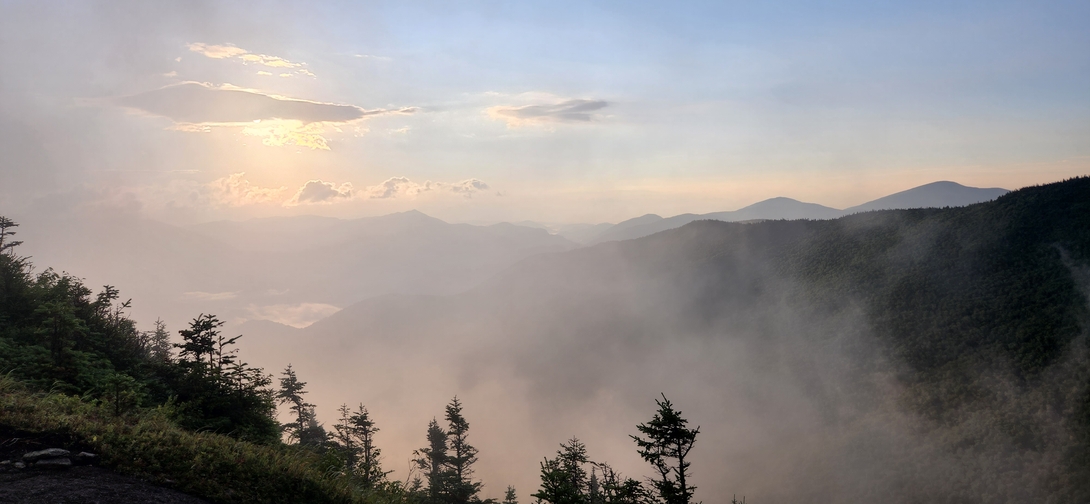 The rising sun shines through clouds above mist-filled mountain valleys viewed from Cascade Mountain.