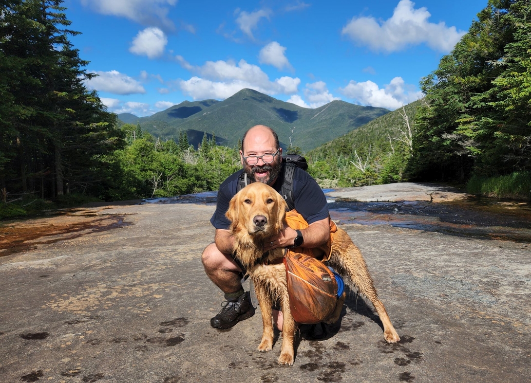 Adam kneels with Sunny at Indian Falls, with Algonquin Peak rising beneath a blue sky behind them.