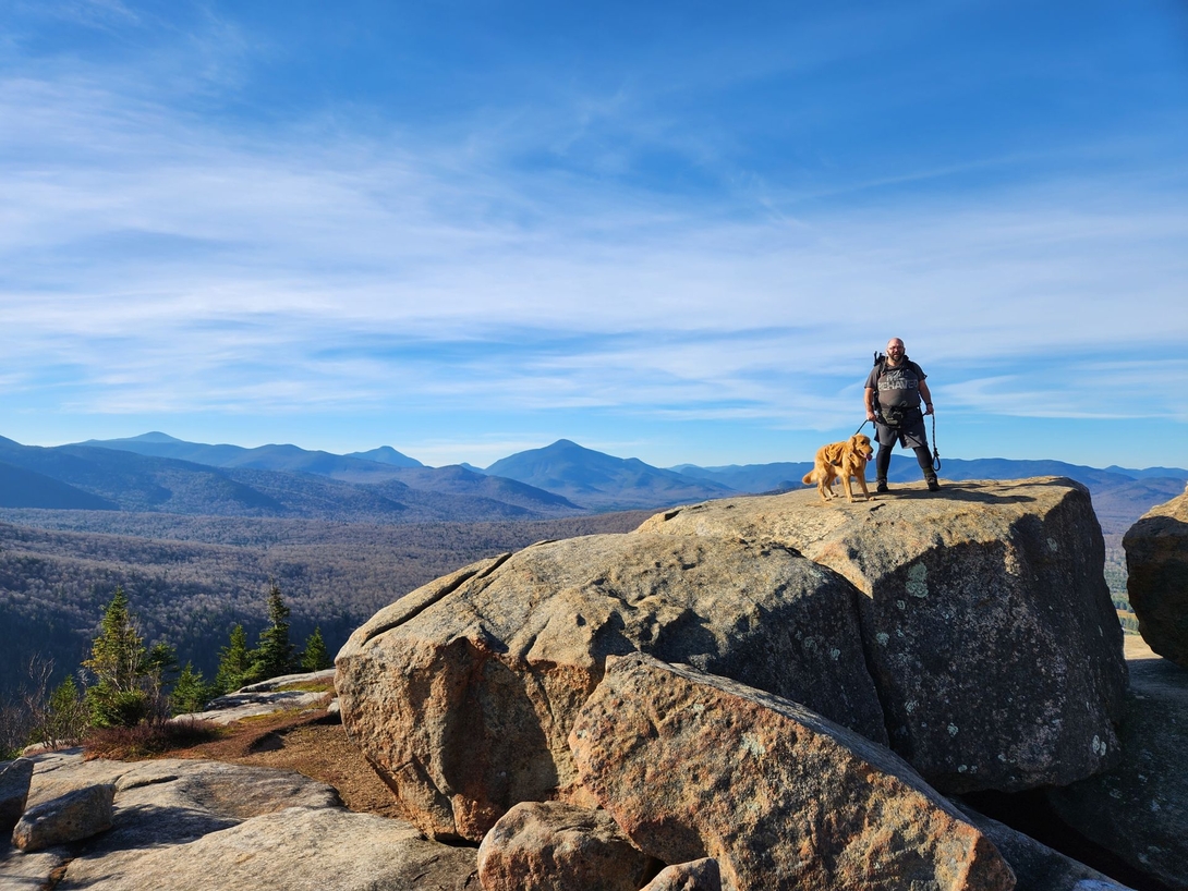 Adam and Sunny stand atop a massive boulder at Balanced Rocks with the High Peaks behind them.