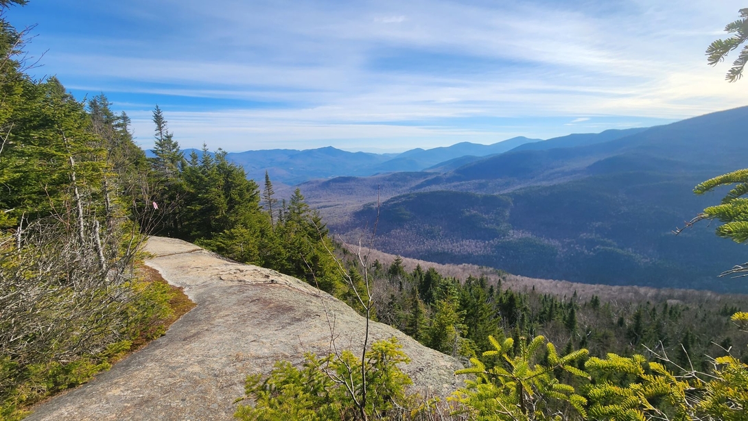 A bare rock ledge at Pitchoff’s summit overlooks the forested valley and neighboring mountains.