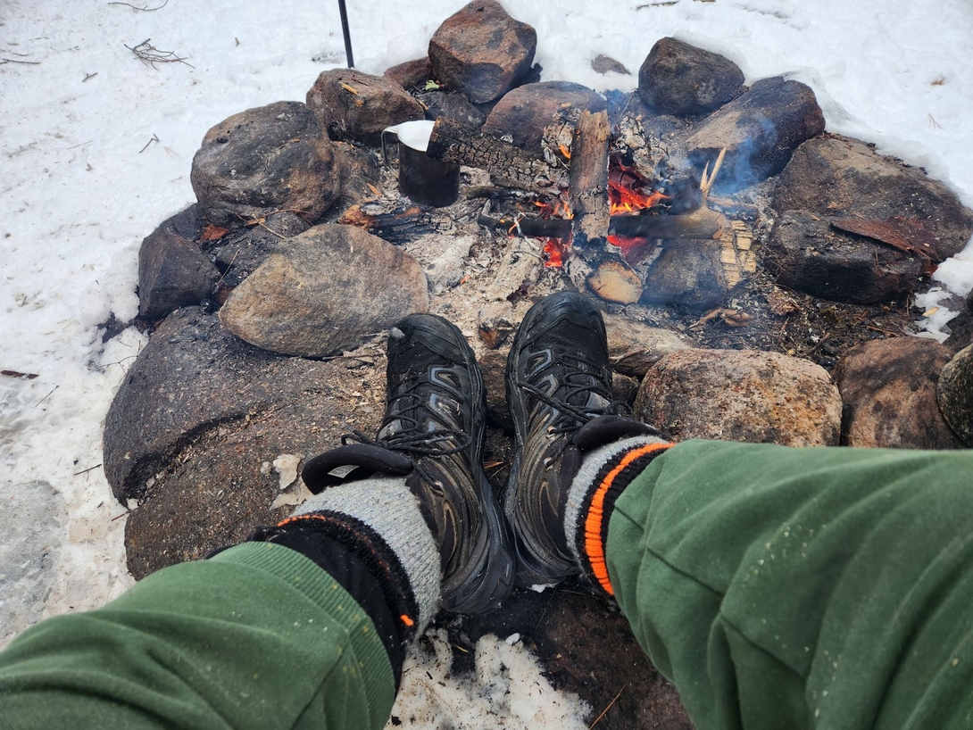 Adam warms his boots beside a small campfire while a metal mug heats over the coals.