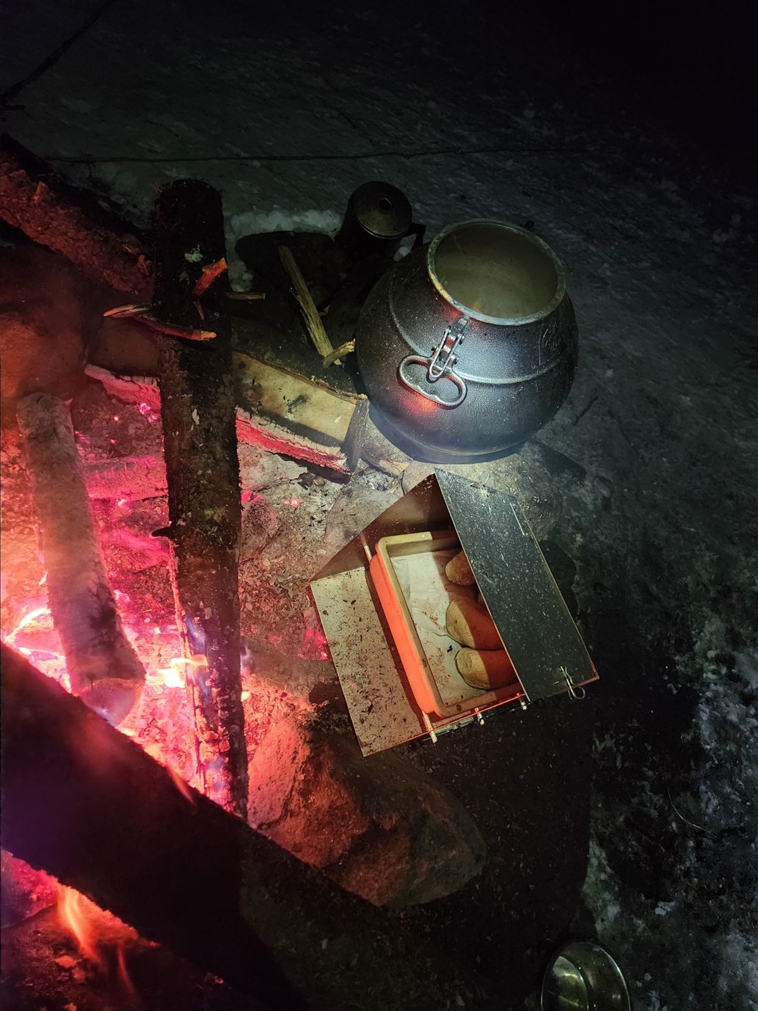 A pressure cooker and camp oven prepare dinner beside the fire, with browned rolls visible inside.