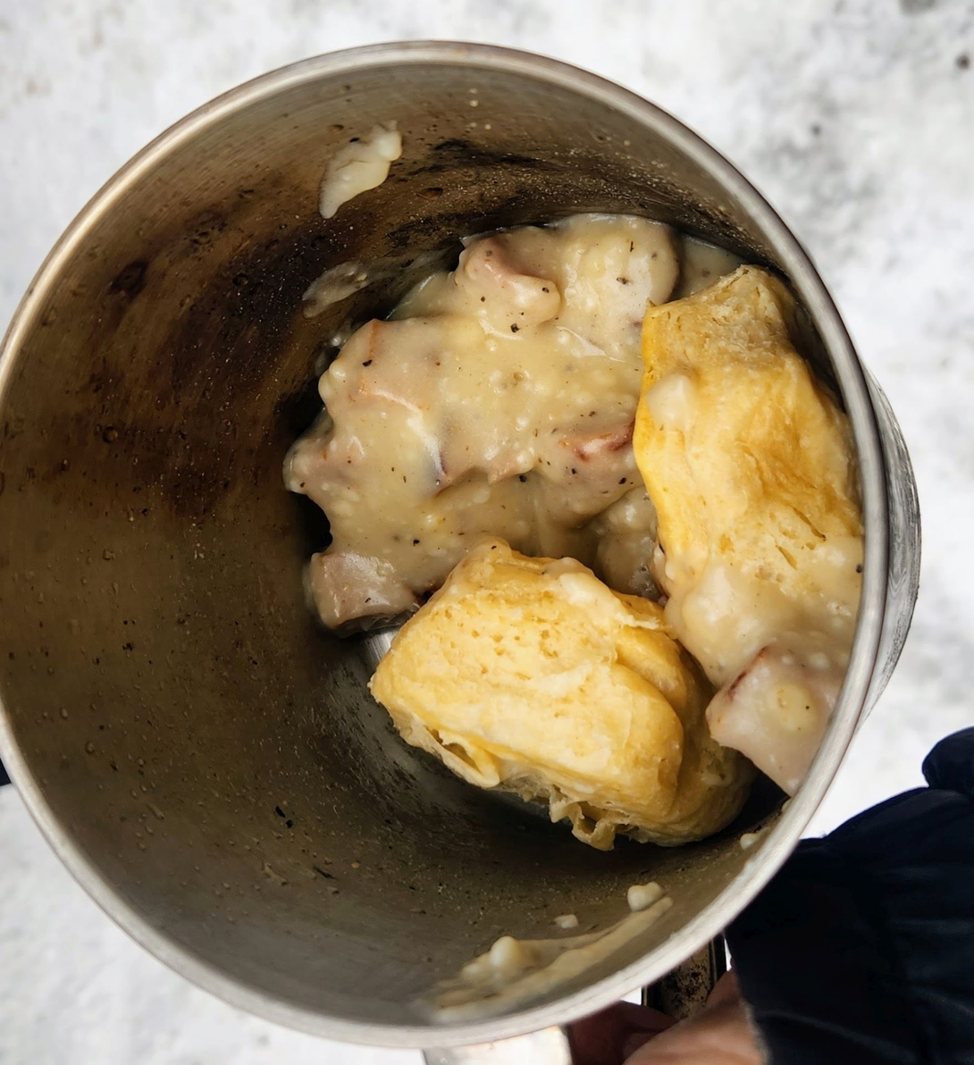 Biscuits covered in sausage gravy fill a metal camping cup at breakfast.