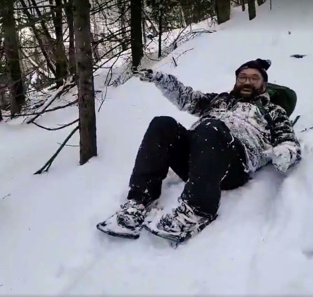 Adam laughs while sliding downhill through deep snow with his snowshoes extended in front of him.